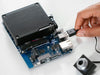 Close-up of a hand plugging a USB cable into the Arduino VENTUNO Q board, showing the heatsink, USB and Ethernet ports, and a camera module nearby.