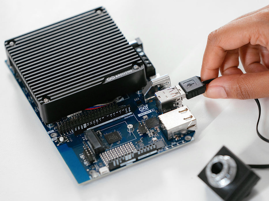 Close-up of a hand plugging a USB cable into the Arduino VENTUNO Q board, showing the heatsink, USB and Ethernet ports, and a camera module nearby.