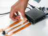 Close-up of a hand attaching an orange flex cable connector to the Arduino VENTUNO Q board, showing the heatsink, USB port, and camera module beside it.