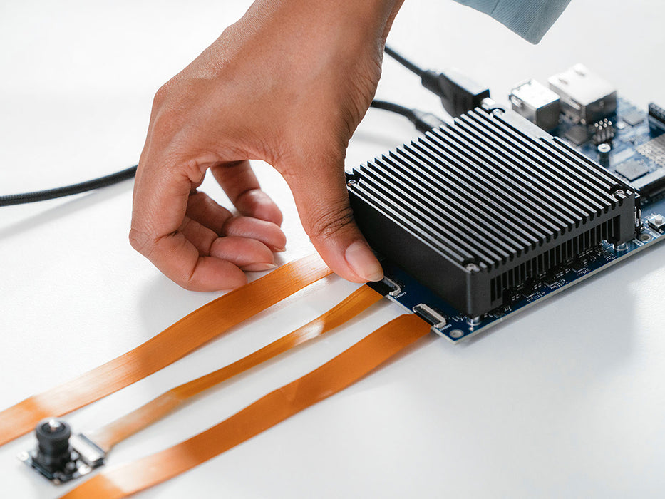Close-up of a hand attaching an orange flex cable connector to the Arduino VENTUNO Q board, showing the heatsink, USB port, and camera module beside it.