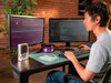 Context photo of a person working at a dual-monitor desk with the compact Arduino VENTUNO Q board sitting on the desk beside a 3D printer filament spool.