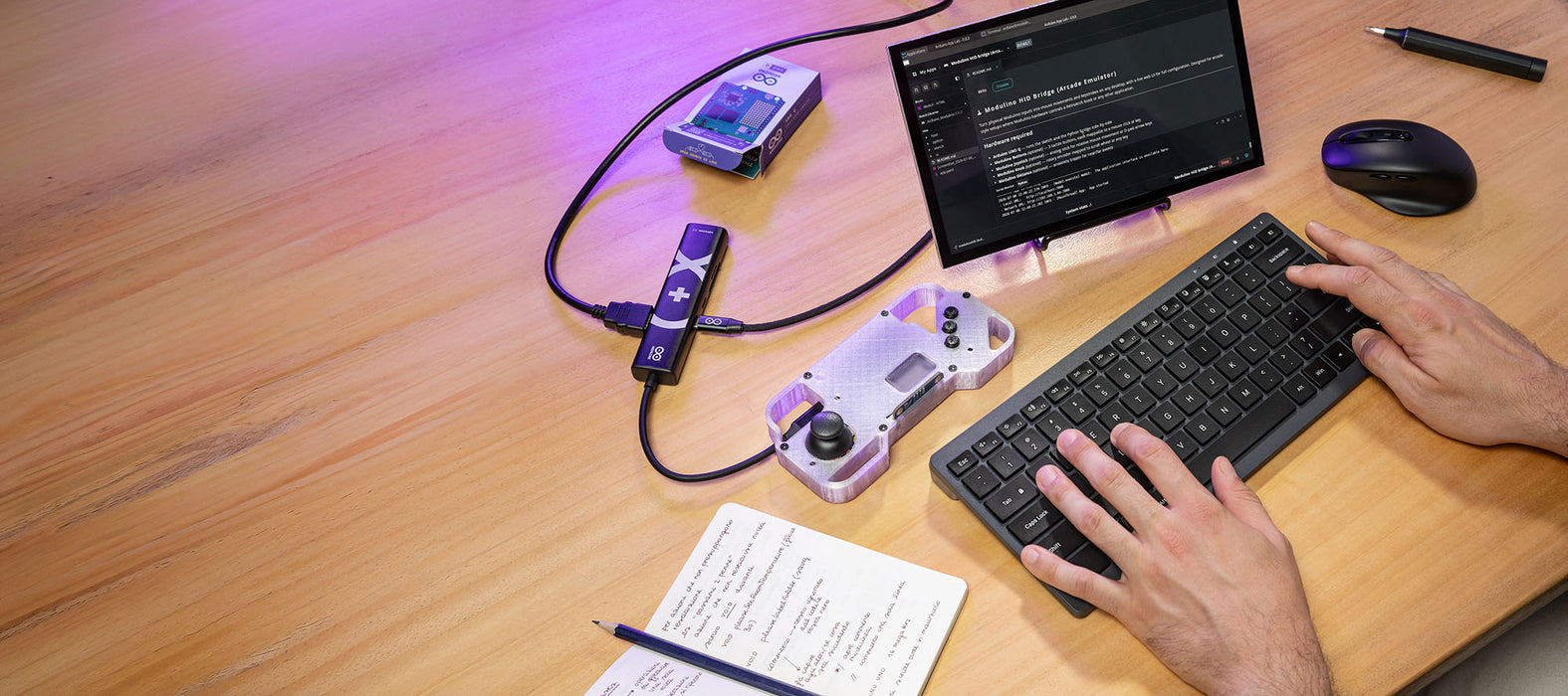 High-angle lifestyle photograph of an organized electronics workstation showcasing a completed DIY project from the Arduino UNO Q Arcade Bundle. A person's hands are typing on a grey keyboard. On the light wood desk, a custom, 3D-printed handheld game controller enclosure—integrating the UNO Q board, joystick, and buttons—is connected via USB-C to an Arduino-branded USB-C hub and a small external monitor displaying project code. A notebook with notes, a pencil, and a mouse complete the setup.
