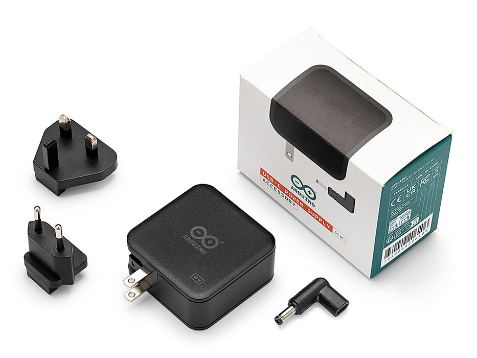 Components of the Arduino 65W USB-C power supply accessory kit, including the power brick, interchangeable US, UK, and EU plugs, and a DC jack adapter, spread out next to its retail box.